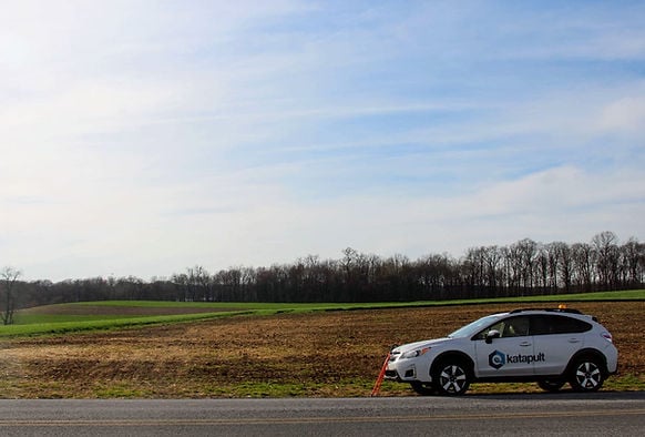 White Subaru with Katapult logo parked near a field 